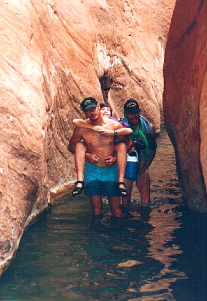 Don carrying Andy through a narrow spot in the canyon with solid sandstone walls and a storm rising. Bill with his pants around his neck